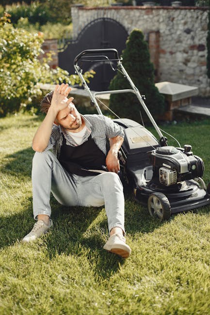 A man with a neatly trimmed beard and short dark hair, dressed in a checked shirt, white trousers, and a black apron, is crouching on a well-maintained lawn in a small yard, holding a pair of gardenin