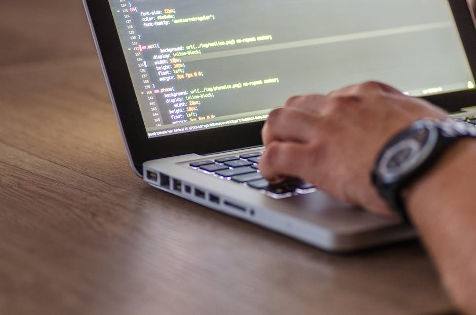A close-up image of a person's hand operating a laptop computer placed on a wooden surface. The laptop screen displays lines of code in a text editor, suggesting programming or web development activit